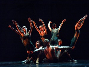 Dancers in a circle, some on the floor holding the ones standing as they lean out of the circle arms extended above their heads