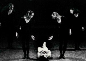 Four dancers in black standing gesture to dancer in white laying on floor