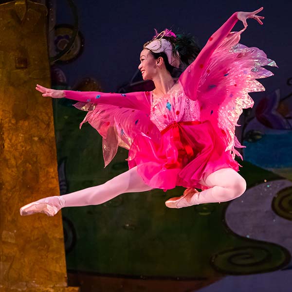 Dancer wearing a short pink dress with sparkly iridescent wings, leaping to the left.