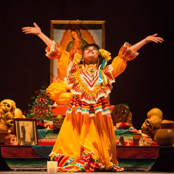 Dancer in a traditional Mexican folklorico dress in front of an ofrenda looking and reaching up.