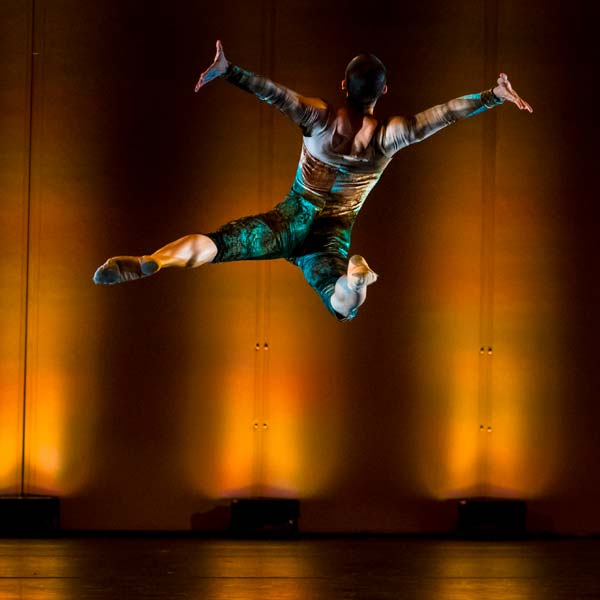 Dancer with back to viewer, wearing green shorts and gold long sleeve. Jumping with arm extended to the side, hands flexed back, right leg bent up left leg extended.