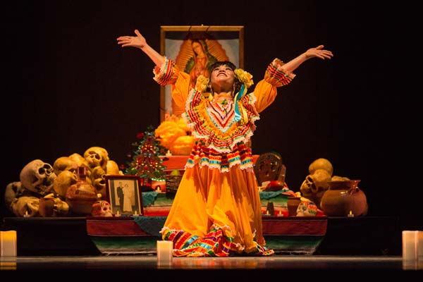 Dancer kneeling down facing front with arms up high in front of ofrenda.