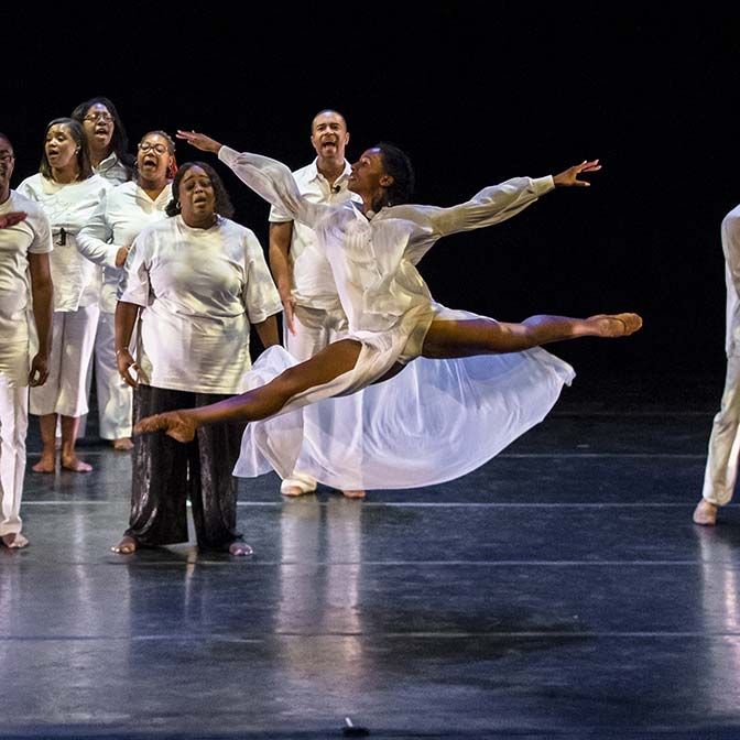 Dancer performing leap to the left in front of choir.