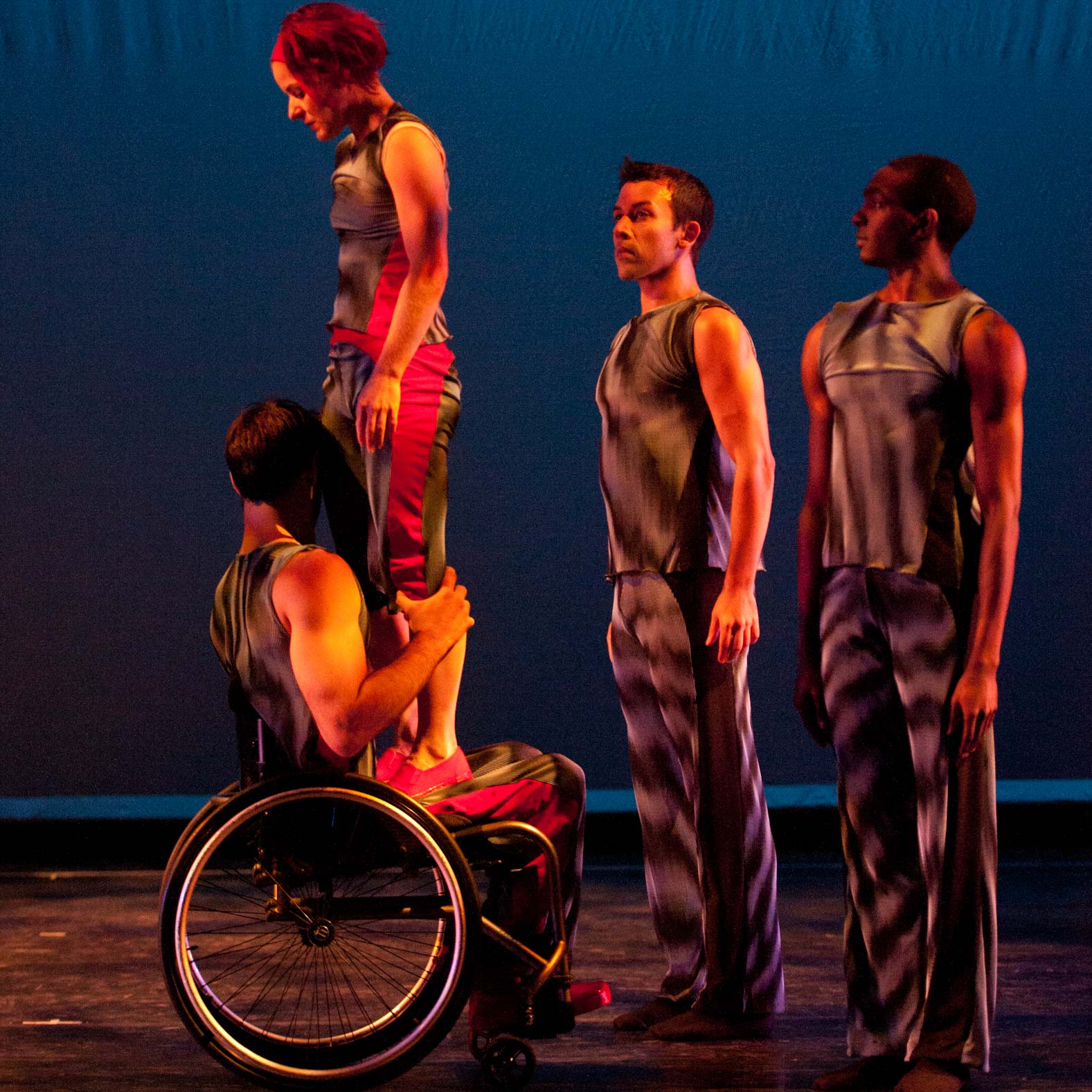 One dancer in wheelchair supporting another dancer while two others stand to right.