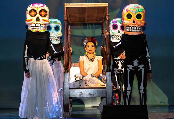 Four dancers in Calavera masks accompanying a dancer dressed as Frida Kahlo on a rolling palanquin.