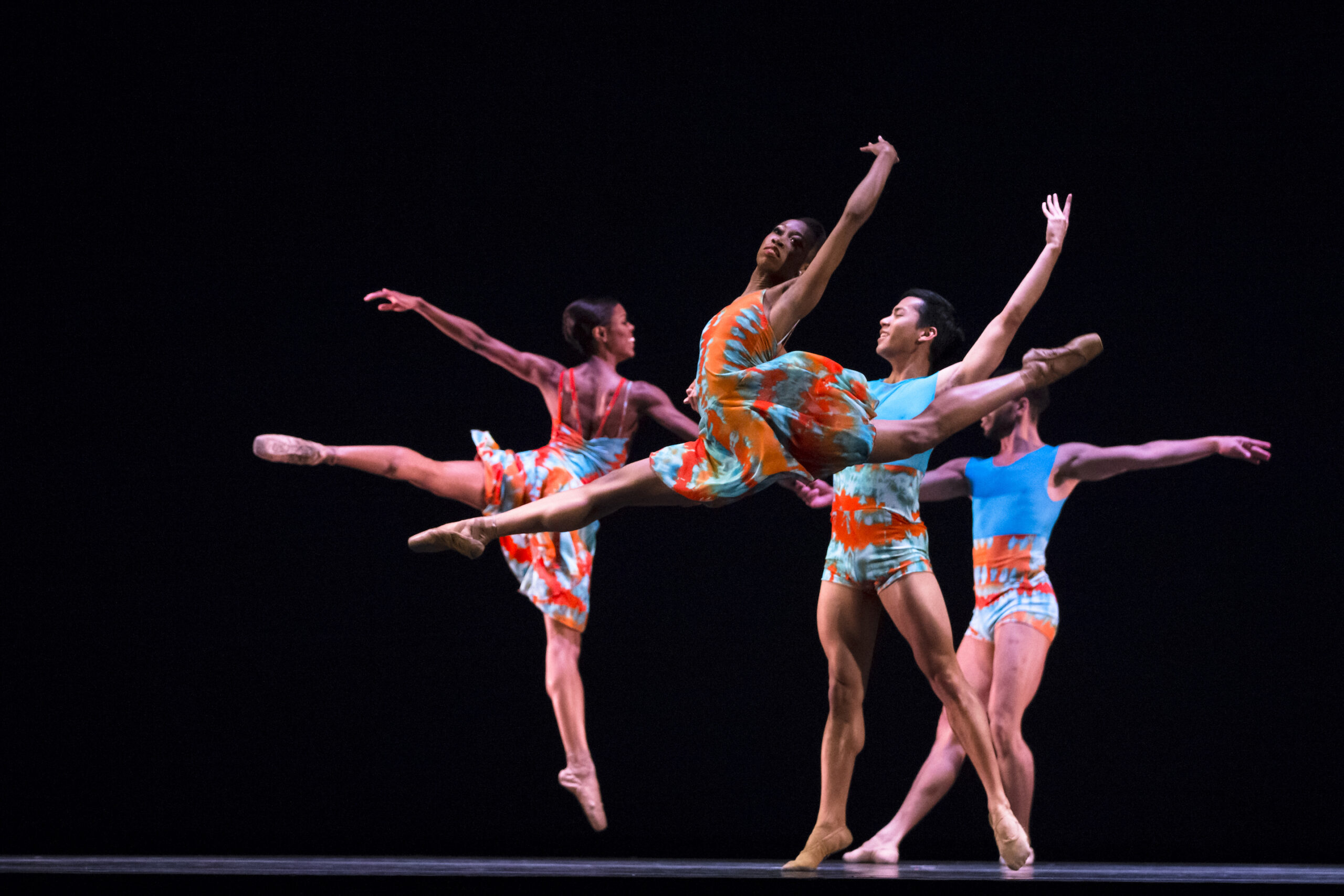 Dancers in blue and orange dancewear perform leaps and other jumps.