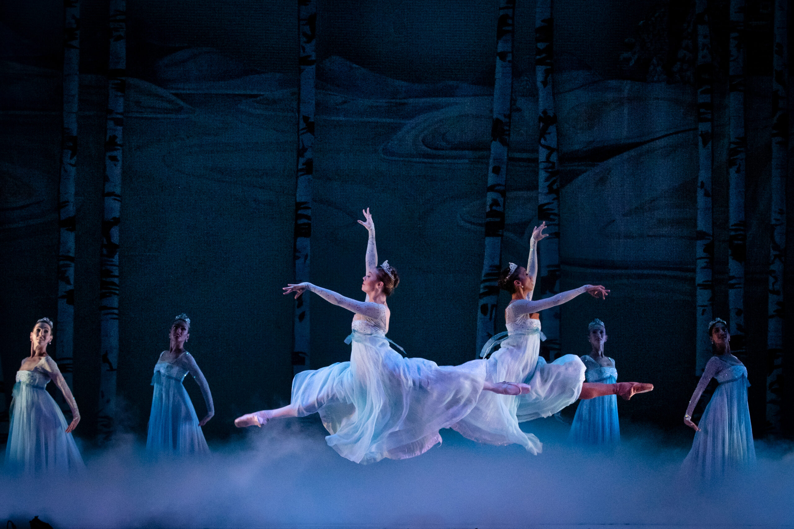 Two dancers dressed in white, flowing gowns perform a leap over mist.