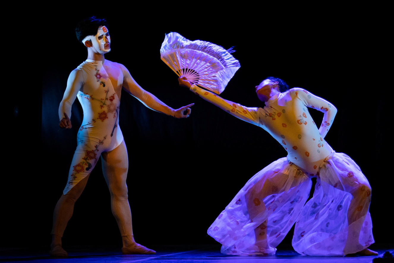 Dancer on left wearing a mask watching dancer on right holding a fan