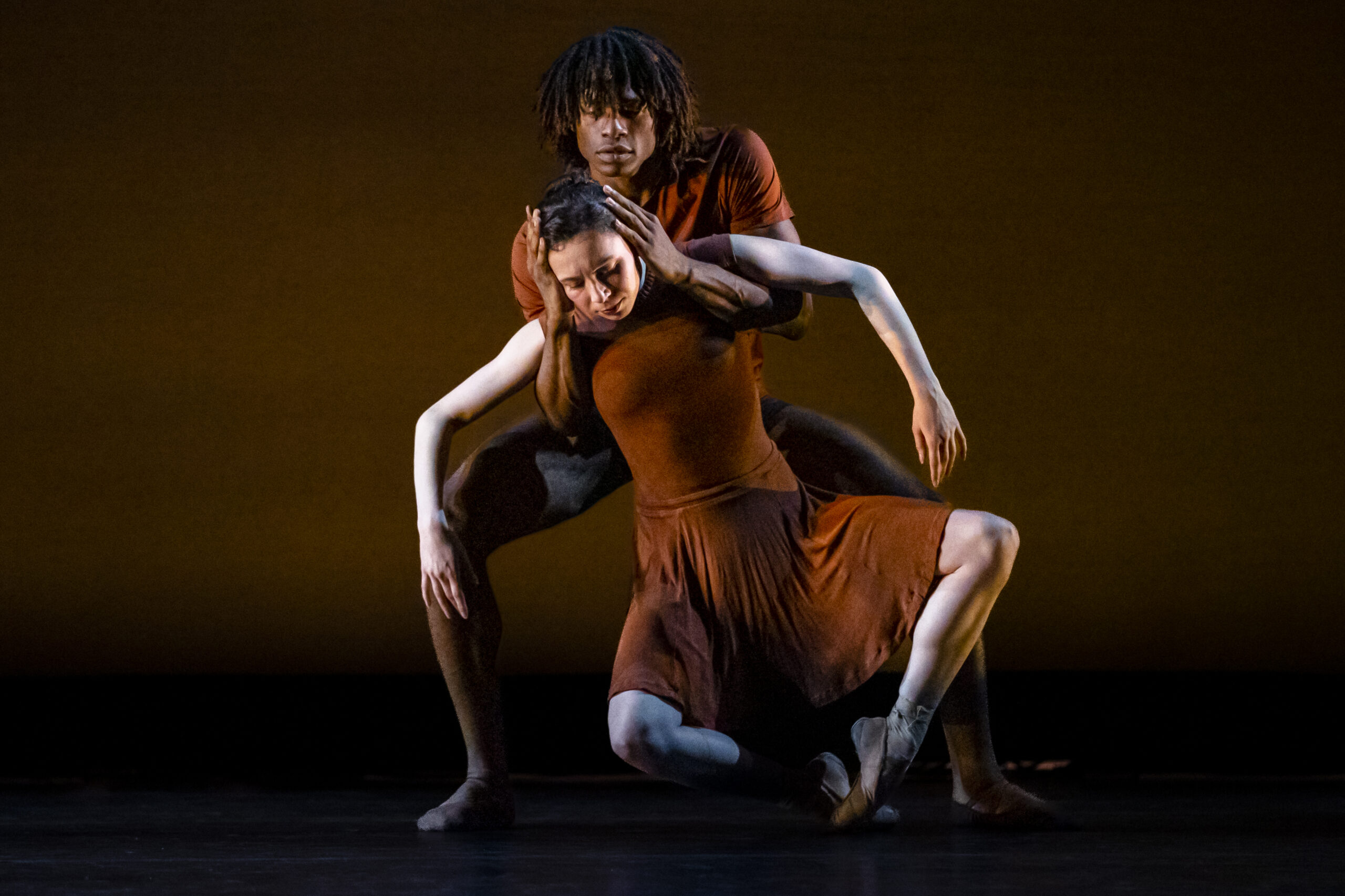 A duet of OBC Dancers in brown costumes dancing low to the ground.