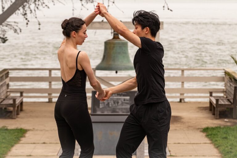 Dancers Jazmine & Lawrence hold hands appearing to perform a turn in front of a bell on Angel Island.